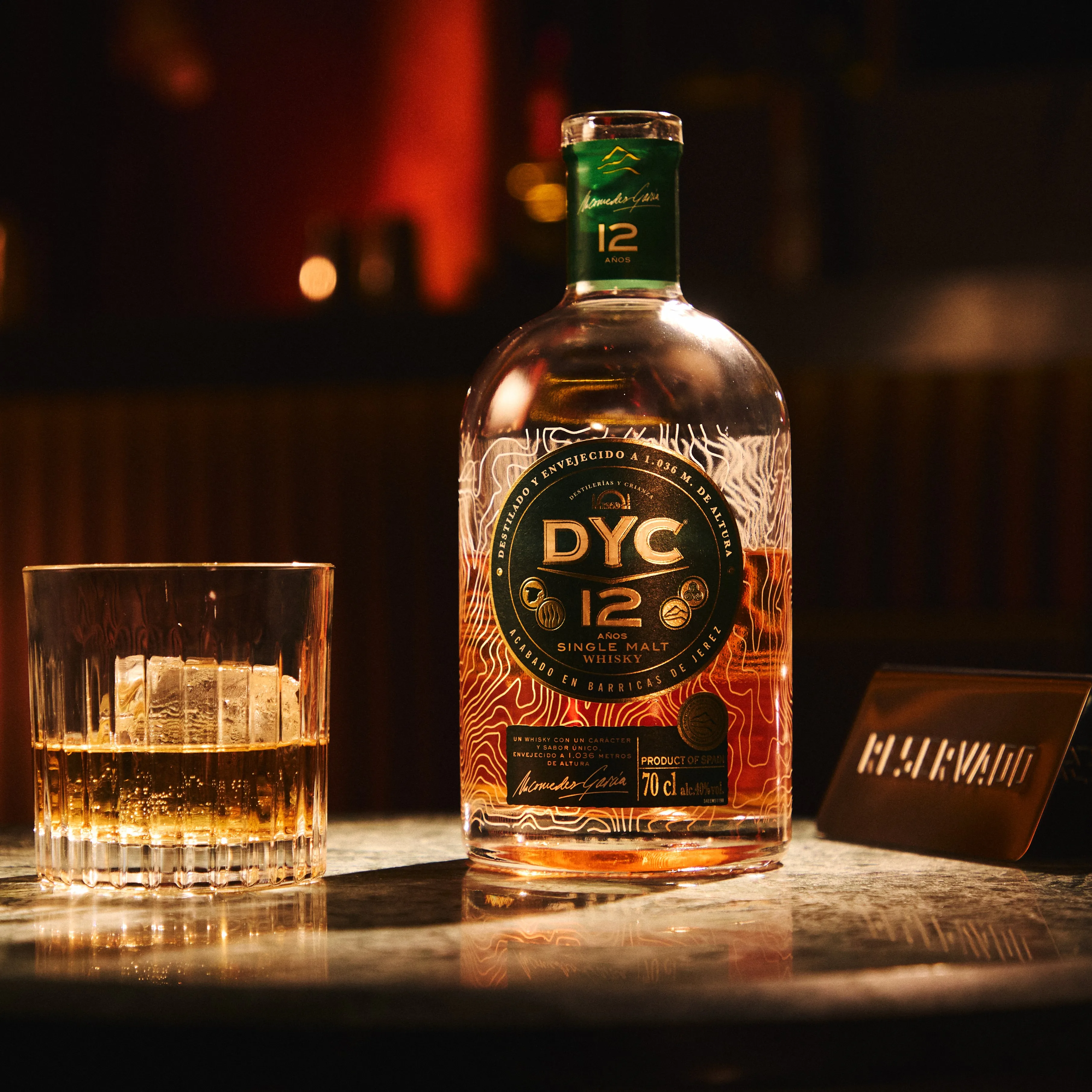 A bottle of DYC 12-year-old single malt whisky stands next to a glass filled with whisky and ice on a dimly lit table.