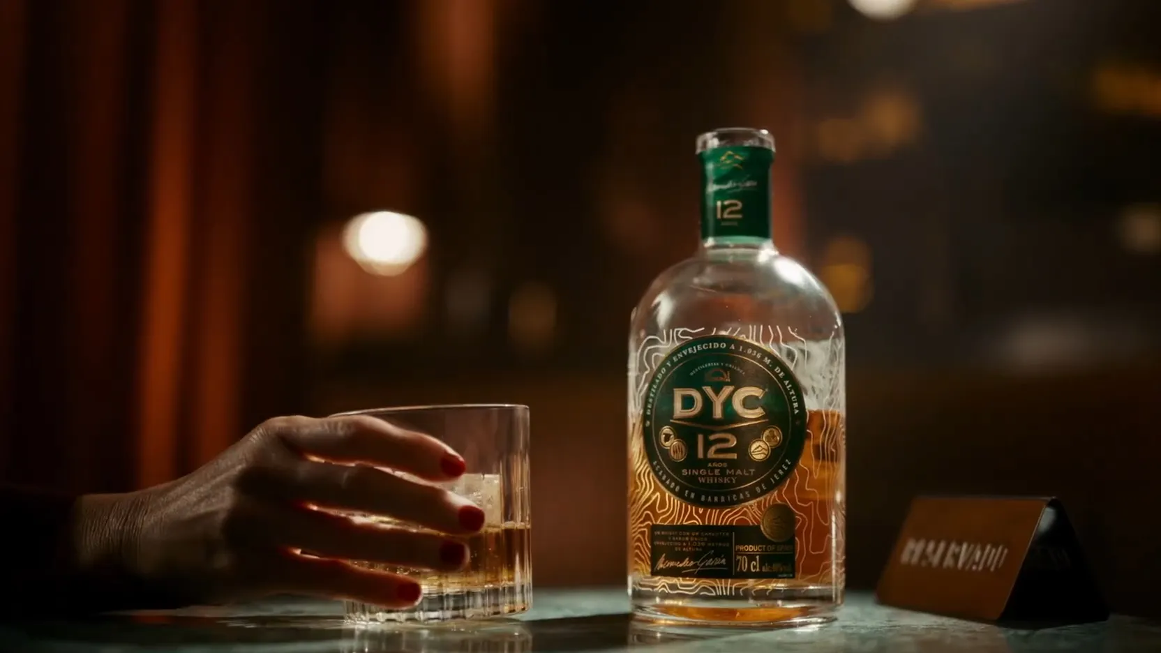 A hand with red nails holds a glass of whisky next to a DYC 12 Single Malt bottle, set against a warm, atmospheric background.
